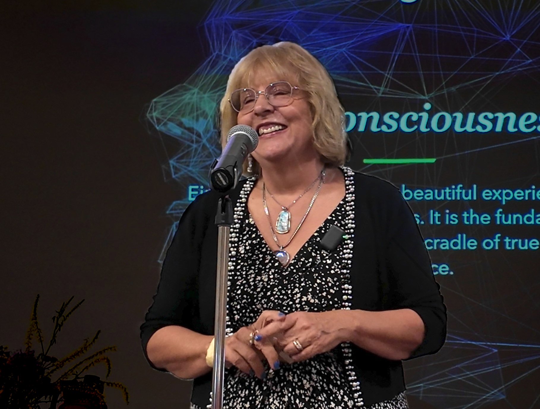 MonaLisa speaking to an audience in front of a projection screen with a graphic about Consciousness behind her.